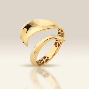 May include: A gold ring with a distinctive open design. The ring has a curved, overlapping band with a smooth, polished finish. The lower band features a geometric pattern. The ring is displayed on a reflective surface.