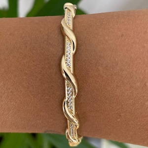 May include: A gold bangle bracelet with a twisted design. The bracelet has a central band of small, clear stones. The bracelet is shown on a person's arm.