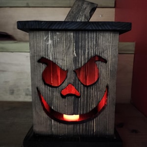 Pode incluir: Uma lanterna de Halloween de madeira com um rosto de abóbora esculpido. A lanterna tem uma forma retangular com uma parte superior e base pretas. Os olhos, o nariz e a boca são recortados, revelando um brilho laranja quente do interior. A madeira tem uma aparência desgastada e rústica.