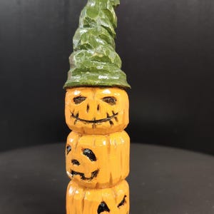 May include: A carved wooden Halloween decoration featuring three stacked pumpkins with carved faces and a green witch hat. The pumpkins are orange with black facial details. The hat is green and textured.