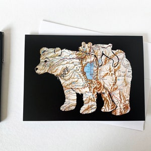 May include: A black card with a brown bear and a smaller brown bear cub on top of it. The bears are made of a map with brown, white, and blue colors. The background is black.