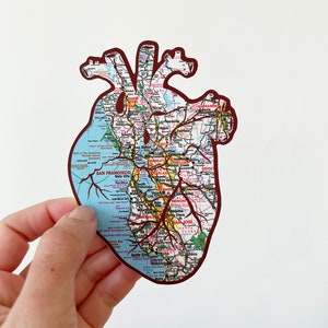 May include: A red heart-shaped sticker with a map of the San Francisco Bay Area printed on it. The map shows major cities and highways. The sticker is held in a hand.