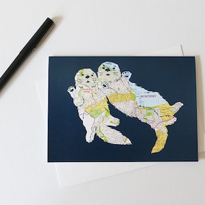 May include: A greeting card with a navy blue background featuring two sea otters made from a map of Monterey, California. The otters are holding hands and the map shows the cities of Pacific Grove and Monterey.