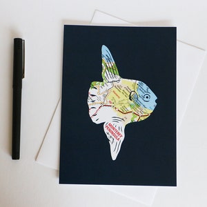 May include: A dark blue card with a white silhouette of a fish made from a map of the Monterey Peninsula. The fish is facing right and has a large eye.