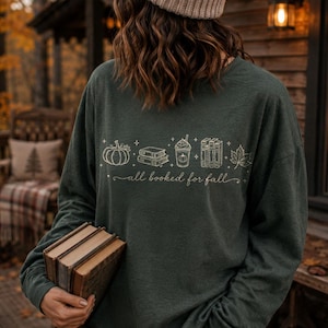 May include: A dark green long-sleeve shirt featuring a white line art graphic of a pumpkin, stacked books, a coffee cup, and a maple leaf, with the text "all booked for fall" written in cursive script across the center of the chest.