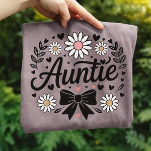 Peut inclure: Un t-shirt mauve avec le mot "Auntie" en écriture cursive, entouré de marguerites, de cœurs et d'un nœud noir. Le design est centré sur le t-shirt, créant une esthétique charmante et féminine.