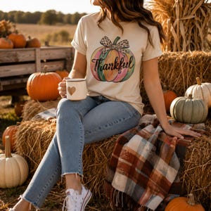 Woman sitting on hay bales wearing a Thankful pumpkin shirt surrounded by pumpkins during the fall harvest season.