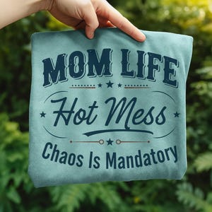 May include: A light blue t-shirt with the words "MOM LIFE Hot Mess Chaos Is Mandatory" in dark blue lettering. The text is arranged in a decorative layout with stars and lines. The shirt is held up by a hand against a blurred green background.
