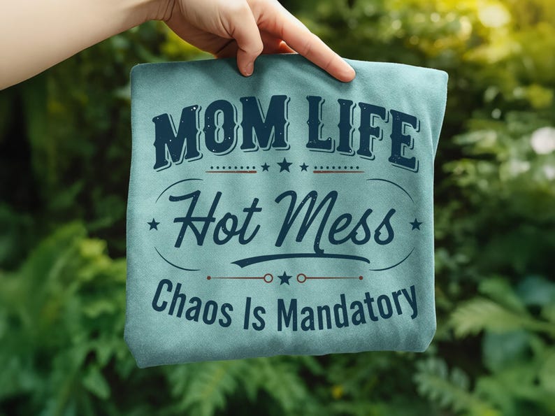 May include: A light blue t-shirt with the words "MOM LIFE Hot Mess Chaos Is Mandatory" in dark blue lettering. The text is arranged in a decorative layout with stars and lines. The shirt is held up by a hand against a blurred green background.