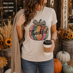 Woman wearing a neutral fall t-shirt featuring a colorful Thankful pumpkin design while holding coffee outside a cozy autumn café.