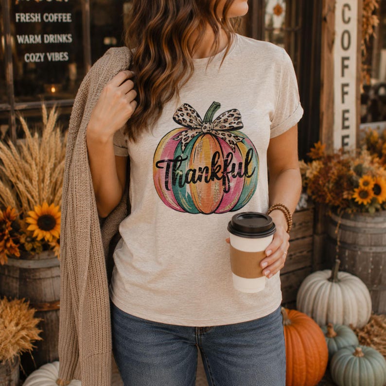 Woman wearing a neutral fall t-shirt featuring a colorful Thankful pumpkin design while holding coffee outside a cozy autumn café.