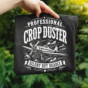 May include: A black t-shirt featuring a white graphic of a vintage propeller airplane. The text reads PROFESSIONAL CROP DUSTER at the top and SILENT BUT DEADLY on a banner at the bottom, with small stars accenting the design.