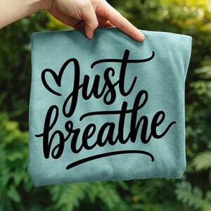 May include: A light teal-colored t-shirt with the words "Just breathe" in black cursive script. The word "Just" has a heart symbol above it. The shirt is held up against a blurred green background.