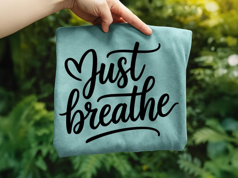 May include: A light teal-colored t-shirt with the words "Just breathe" in black cursive script. The word "Just" has a heart symbol above it. The shirt is held up against a blurred green background.