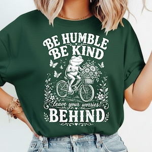 Puede incluir: Una camiseta verde oscuro con un estampado gráfico blanco de una rana montando una bicicleta con una cesta de flores. El texto dice BE HUMBLE BE KIND leave your worries BEHIND, rodeado de pequeñas mariposas y motivos florales.