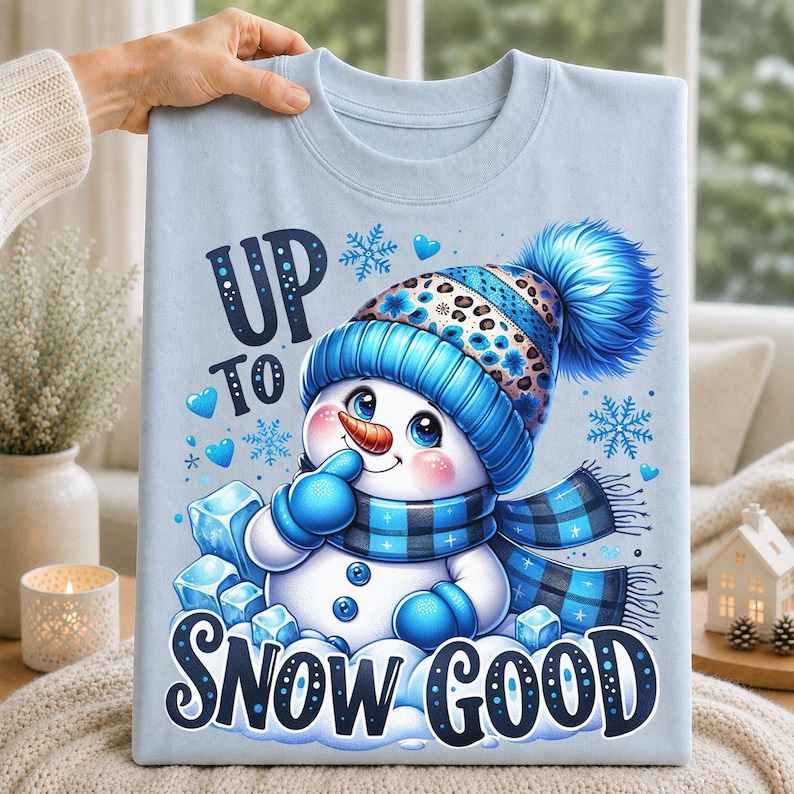 Blue t-shirt featuring a cute snowman wearing a blue pom-pom hat and plaid scarf with Up to Snow Good winter lettering and snowflakes