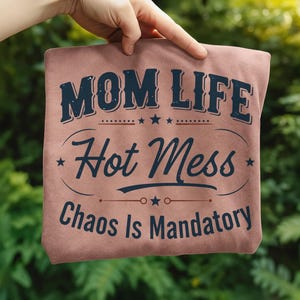 May include: A dusty rose-colored t-shirt with the text "MOM LIFE Hot Mess Chaos Is Mandatory" in navy blue. The text is arranged in a stylized design with stars and decorative elements.