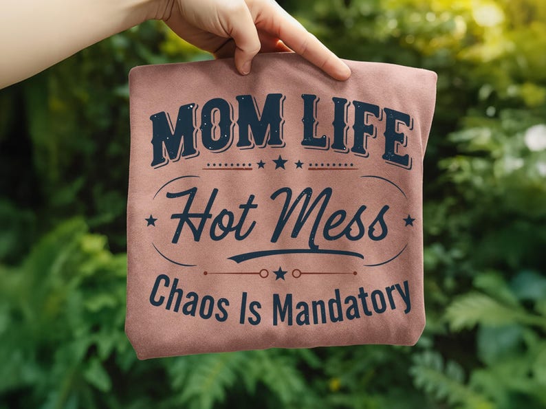 May include: A dusty rose-colored t-shirt with the text "MOM LIFE Hot Mess Chaos Is Mandatory" in navy blue. The text is arranged in a stylized design with stars and decorative elements.