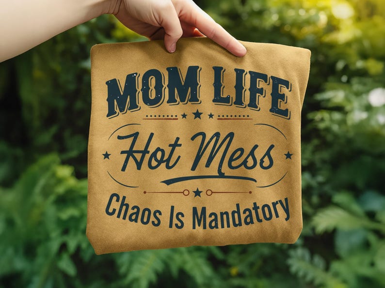 May include: A tan t-shirt with the words "MOM LIFE Hot Mess Chaos Is Mandatory" in navy blue lettering. The text is arranged in a vintage style with stars and decorative elements. The shirt is held up against a blurred green background.