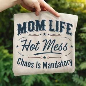 May include: A beige tote bag with the text "MOM LIFE Hot Mess Chaos Is Mandatory" in navy blue. The text is arranged in a vintage-style design with stars and decorative lines. The bag is held up against a blurred green background.