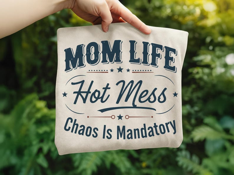 May include: A beige tote bag with the text "MOM LIFE Hot Mess Chaos Is Mandatory" in navy blue. The text is arranged in a vintage-style design with stars and decorative lines. The bag is held up against a blurred green background.