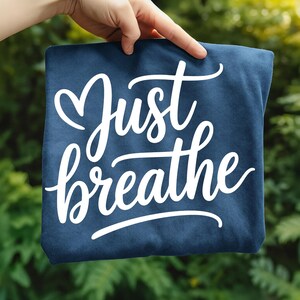 May include: A navy blue square textile with the words "Just breathe" in white cursive script. The text is centered and the fabric is held up against a blurred green background.