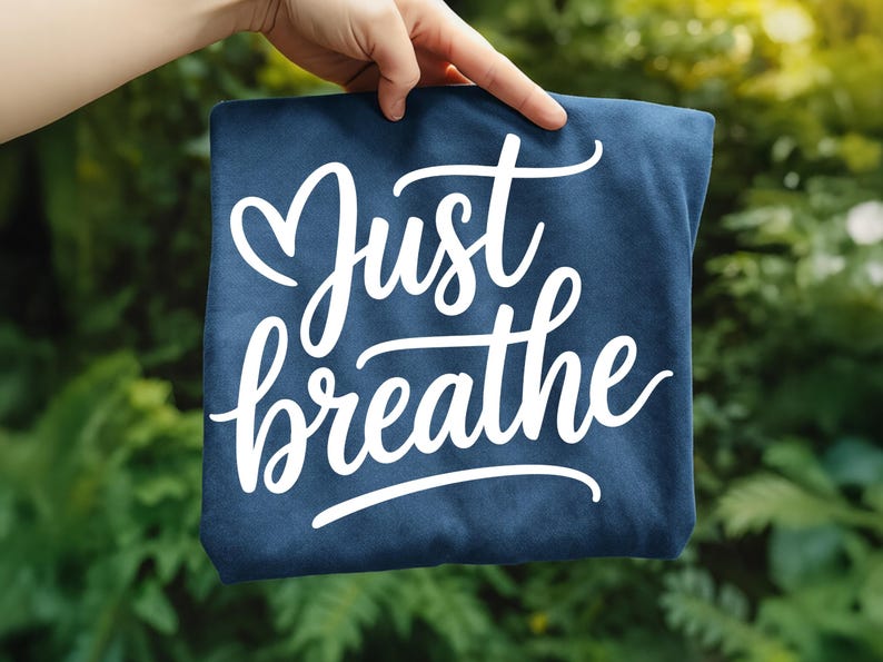 May include: A navy blue square textile with the words "Just breathe" in white cursive script. The text is centered and the fabric is held up against a blurred green background.