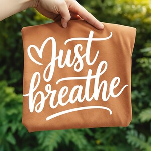 May include: A brown tote bag with the words "Just breathe" in white cursive script. The bag is held up against a backdrop of green foliage.