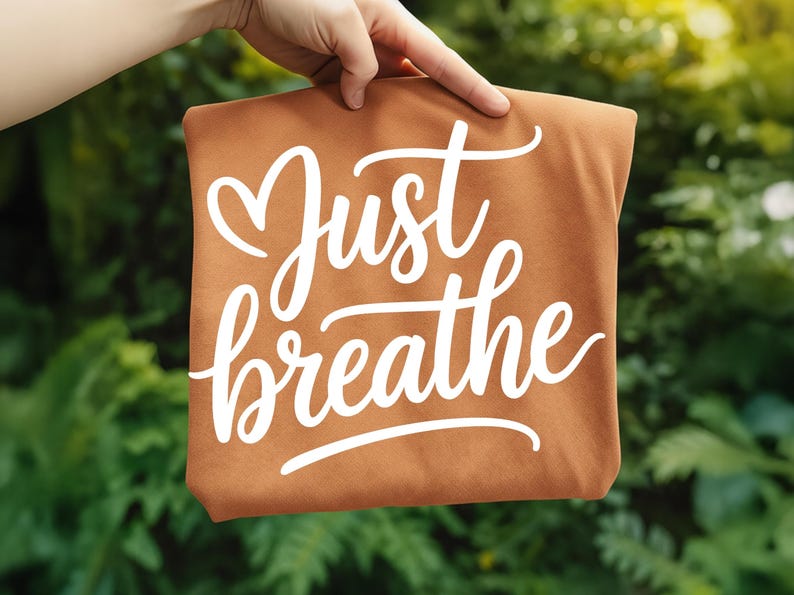 May include: A brown tote bag with the words "Just breathe" in white cursive script. The bag is held up against a backdrop of green foliage.