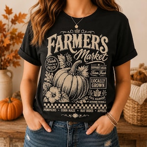 May include: A black t-shirt featuring a cream-colored graphic with a large pumpkin, leaves, and text that reads Farmer's Market, Fresh From The Patch, Support Local, Farm Fresh, Locally Grown, Fall Harvest, Autumn Picked, Pumpkins, Corn Maze, Hay Rides, Apples.