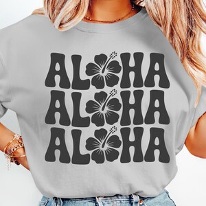 Pode incluir: Camiseta cinza claro com a palavra "ALOHA" repetida tr&ecirc;s vezes em cinza escuro, cada uma com um gr&aacute;fico de flor de hibisco. A camiseta tem gola redonda e mangas curtas, perfeita para um ambiente tropical casual.