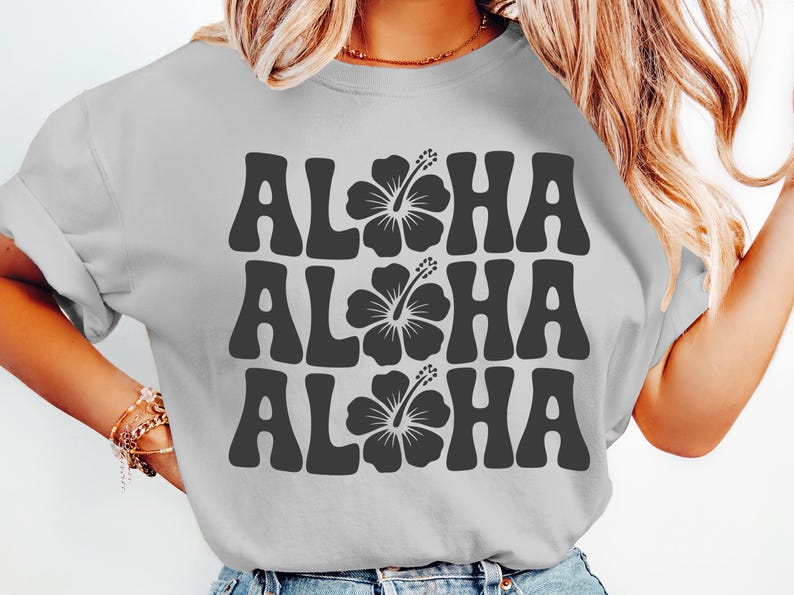 Pode incluir: Camiseta cinza claro com a palavra "ALOHA" repetida tr&ecirc;s vezes em cinza escuro, cada uma com um gr&aacute;fico de flor de hibisco. A camiseta tem gola redonda e mangas curtas, perfeita para um ambiente tropical casual.