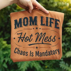 May include: A tan t-shirt with the words "MOM LIFE Hot Mess Chaos Is Mandatory" in dark blue lettering. The text is arranged in a stylized design with stars and decorative lines. The shirt is held up against a green, leafy background.
