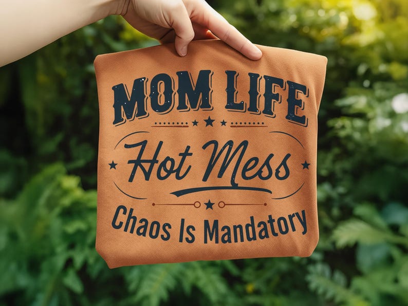 May include: A tan t-shirt with the words "MOM LIFE Hot Mess Chaos Is Mandatory" in dark blue lettering. The text is arranged in a stylized design with stars and decorative lines. The shirt is held up against a green, leafy background.