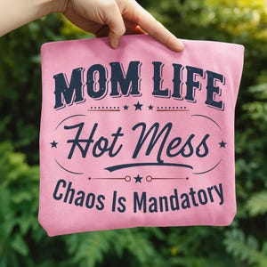 May include: Pink t-shirt with the text "MOM LIFE Hot Mess Chaos Is Mandatory" in navy blue. The design includes stars and decorative lines. The shirt is held up against a blurred green background.