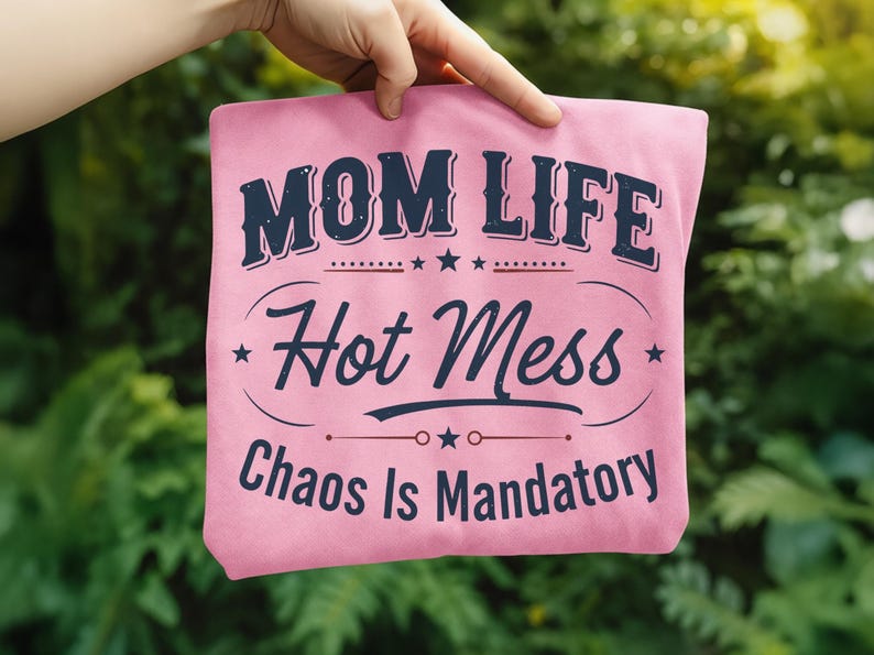 May include: Pink t-shirt with the text "MOM LIFE Hot Mess Chaos Is Mandatory" in navy blue. The design includes stars and decorative lines. The shirt is held up against a blurred green background.