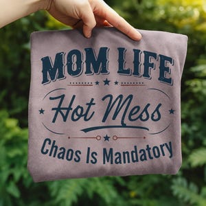 May include: A mauve-colored t-shirt with the words "MOM LIFE Hot Mess Chaos Is Mandatory" in a navy blue, vintage-style font. The text is arranged in a layered design with stars and decorative lines. The shirt is held up by a hand.