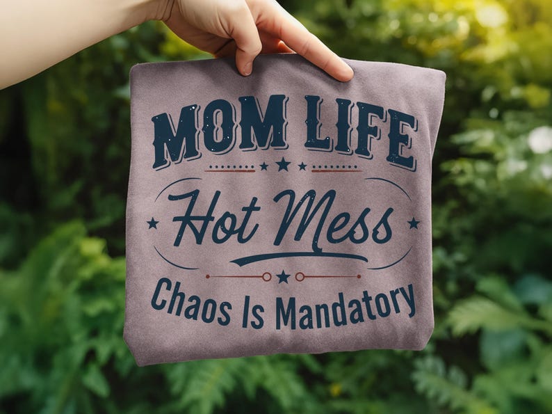 May include: A mauve-colored t-shirt with the words "MOM LIFE Hot Mess Chaos Is Mandatory" in a navy blue, vintage-style font. The text is arranged in a layered design with stars and decorative lines. The shirt is held up by a hand.