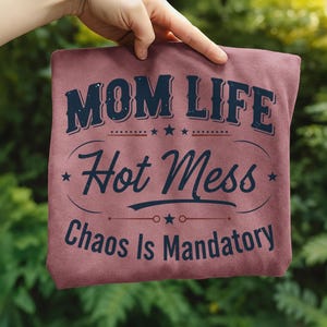 May include: A maroon t-shirt with the words "MOM LIFE Hot Mess Chaos Is Mandatory" in a navy blue, vintage-style font. The text is arranged in a layered design with stars and decorative lines.