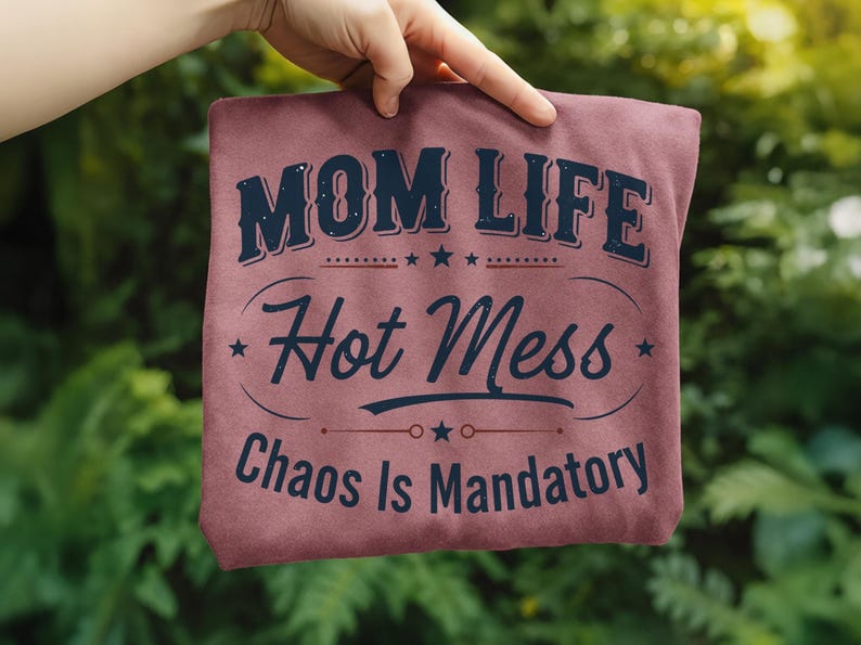 May include: A maroon t-shirt with the words "MOM LIFE Hot Mess Chaos Is Mandatory" in a navy blue, vintage-style font. The text is arranged in a layered design with stars and decorative lines.