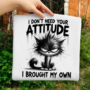 May include: White tote bag featuring a black and white illustration of an angry cat. The text "I DON'T NEED YOUR ATTITUDE I BROUGHT MY OWN" is printed above and below the cat image. The cat has a grumpy expression.