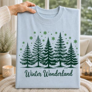 Winter Wonderland snowy forest T-shirt design displayed on a light blue shirt with evergreen trees and snowflakes