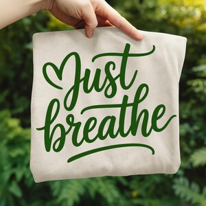 May include: A beige tote bag with the words "Just breathe" in green script. The bag is held up against a blurred green background of foliage.