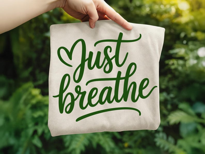 May include: A beige tote bag with the words "Just breathe" in green script. The bag is held up against a blurred green background of foliage.