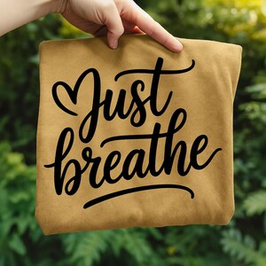 May include: A tan-colored t-shirt with the words "Just breathe" in black cursive script. The word "Just" has a heart shape above it. The shirt is held up by a hand against a blurred green background.