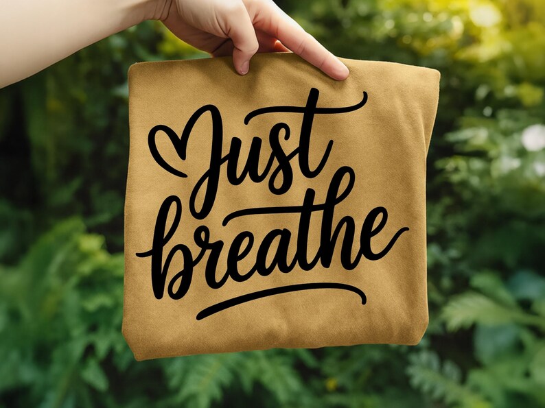 May include: A tan-colored t-shirt with the words "Just breathe" in black cursive script. The word "Just" has a heart shape above it. The shirt is held up by a hand against a blurred green background.