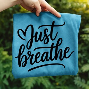 May include: A teal-colored t-shirt with the words "Just breathe" in a black, cursive font. The word "Just" has a heart-shaped design above it. The shirt is held up by a hand against a blurred green background.