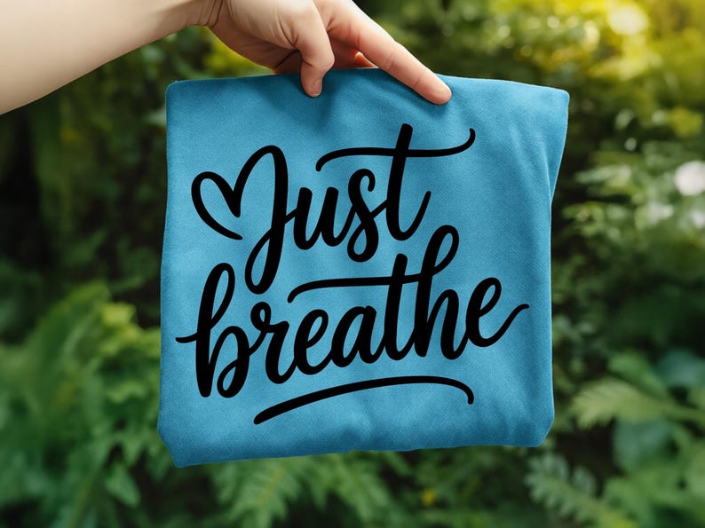 May include: A teal-colored t-shirt with the words "Just breathe" in a black, cursive font. The word "Just" has a heart-shaped design above it. The shirt is held up by a hand against a blurred green background.