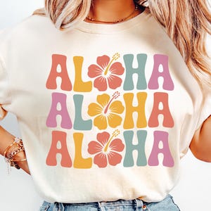 Pode incluir: Camiseta creme com a palavra "ALOHA" em fonte retr&ocirc;, repetida tr&ecirc;s vezes com gr&aacute;ficos de flores de hibisco. As letras s&atilde;o em tons de rosa, laranja, amarelo e azul. Um design casual e de ver&atilde;o.