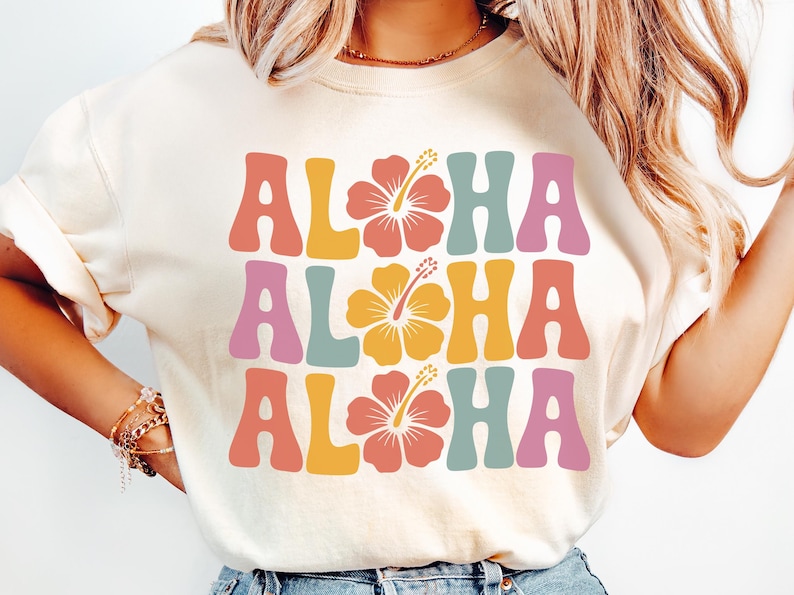 Pode incluir: Camiseta creme com a palavra "ALOHA" em fonte retr&ocirc;, repetida tr&ecirc;s vezes com gr&aacute;ficos de flores de hibisco. As letras s&atilde;o em tons de rosa, laranja, amarelo e azul. Um design casual e de ver&atilde;o.